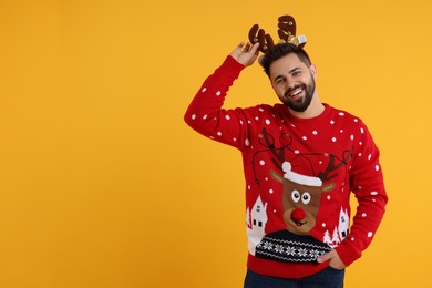 Photo of Happy young man in Christmas sweater and reindeer headband on orange background. Space for text