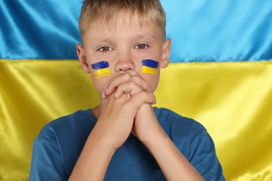 Photo of Little boy with clasped hands praying for Ukraine near national flag. No war concept
