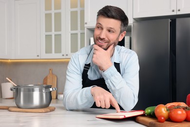 Photo of Adult man reading recipe book at white marble table in kitchen