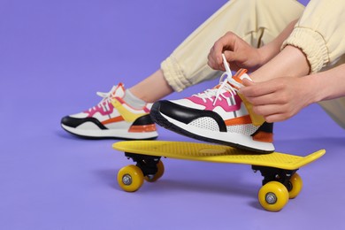 Photo of Woman tying lace of sneaker on skateboard against purple background, closeup