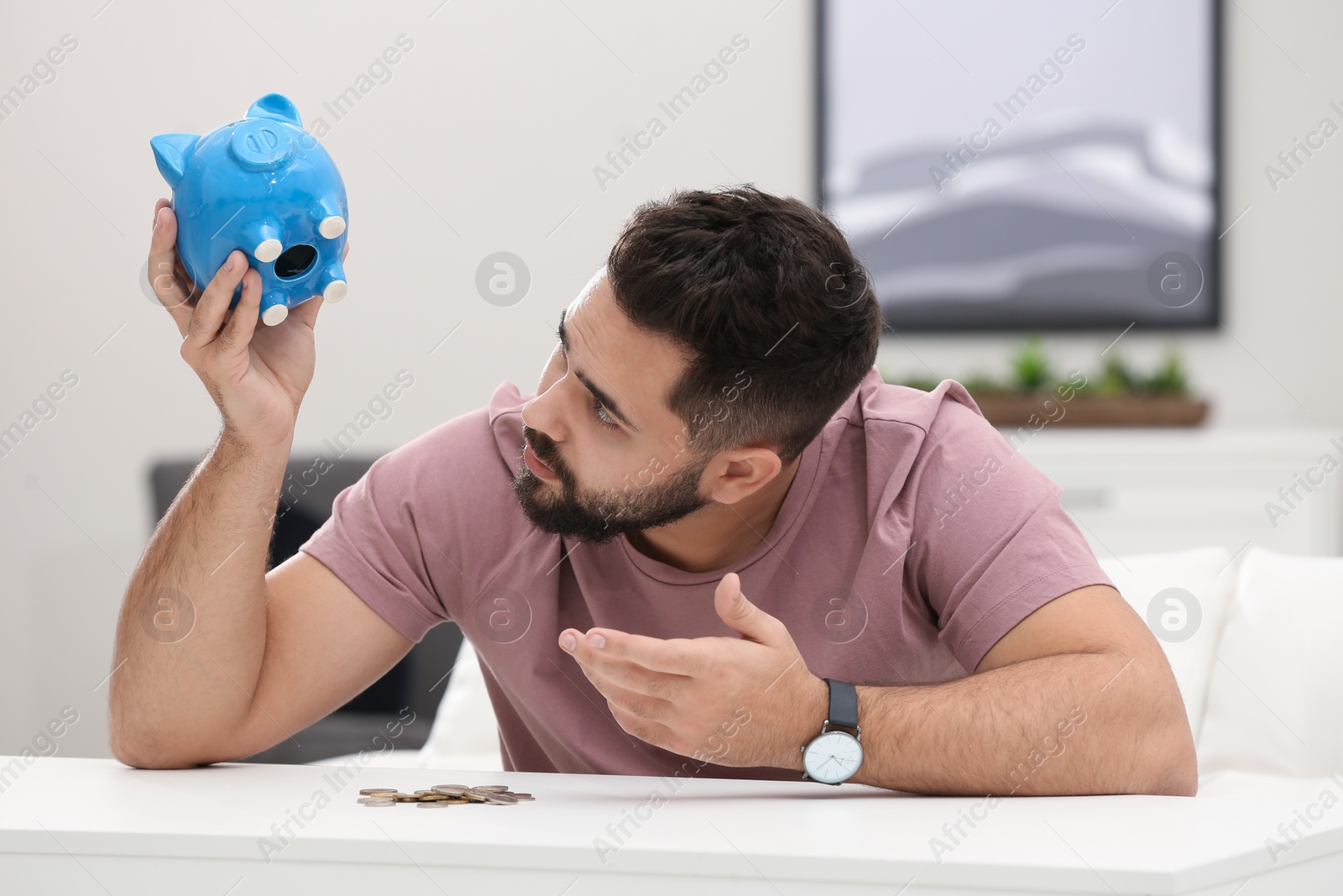 Photo of Worried young man with piggy bank and money at white table indoors