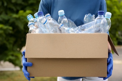 Woman holding cardboard box with used plastic bottles outdoors, closeup. Recycling problem