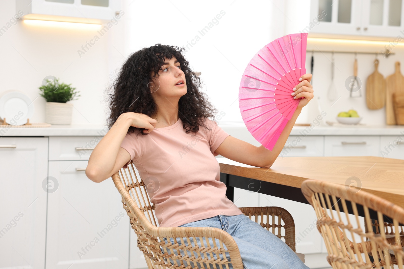 Photo of Young woman waving pink hand fan to cool herself at table in kitchen