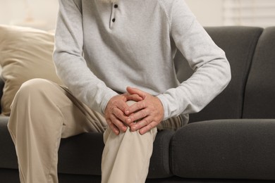 Senior man suffering from knee pain on sofa indoors, closeup. Rheumatism symptom