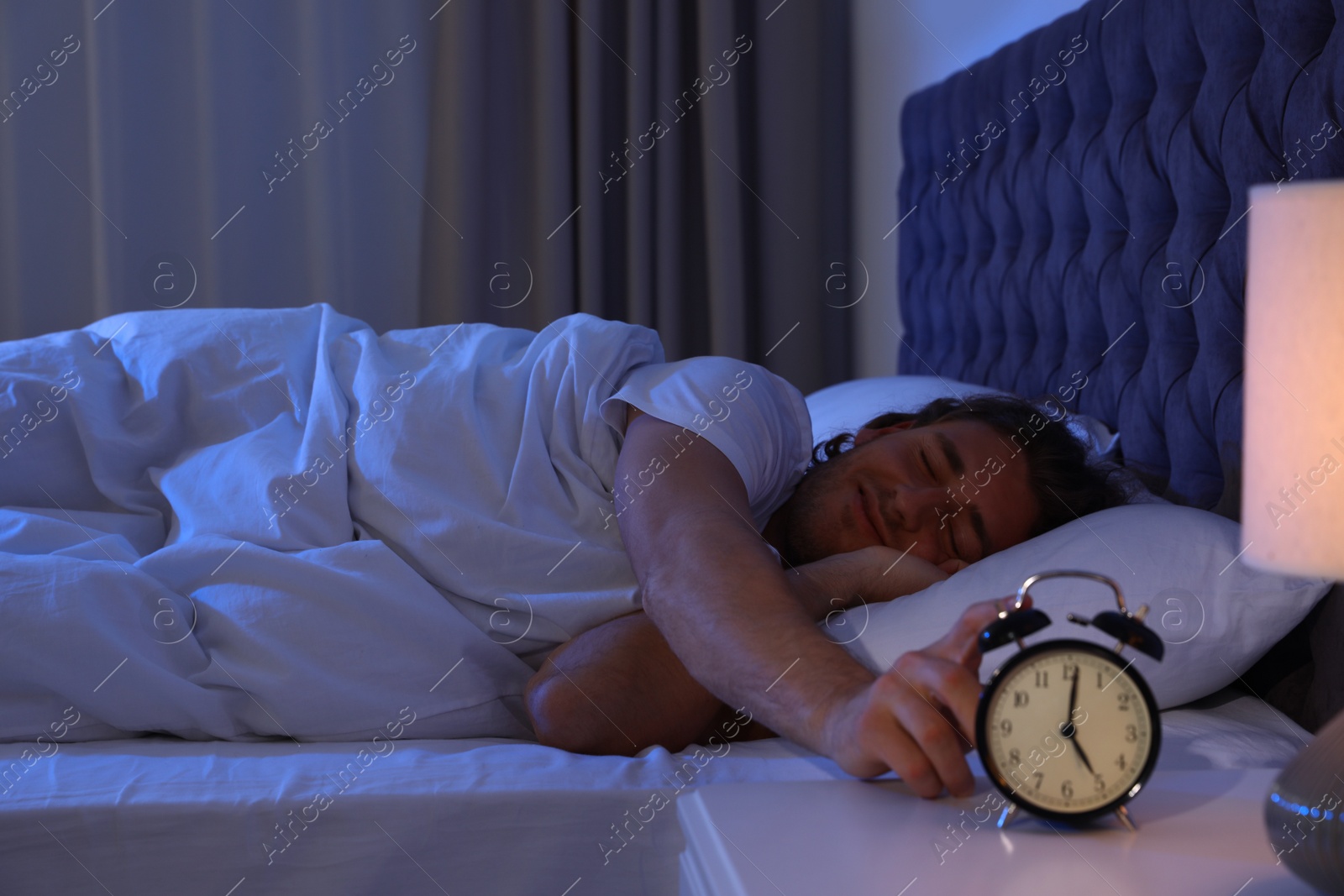 Photo of Sleepy young man turning off alarm clock on nightstand. Bedtime