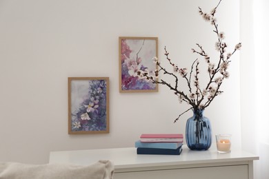 Photo of Flowering tree twigs in glass vase, burning candle and books on table near white wall at home