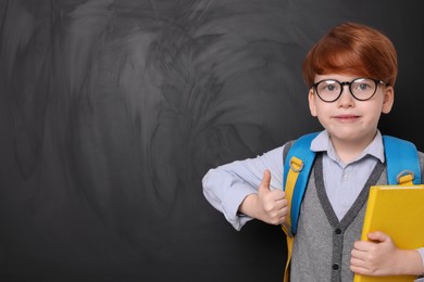 Photo of Cute schoolboy in glasses with book showing thumb up near blackboard. Space for text