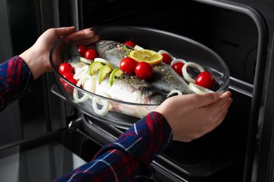 Photo of Woman putting glass baking tray with sea bass fish and ingredients into oven, closeup