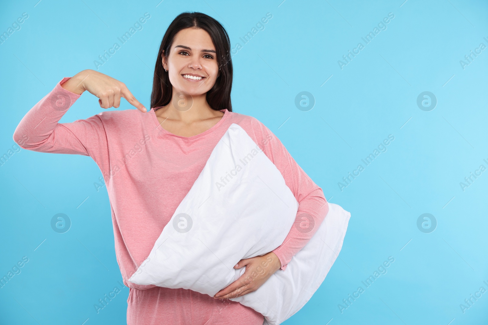 Photo of Happy young woman pointing at soft pillow on light blue background, space for text