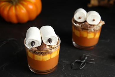 Photo of Glasses with delicious dessert decorated as monsters on black table. Halloween treat