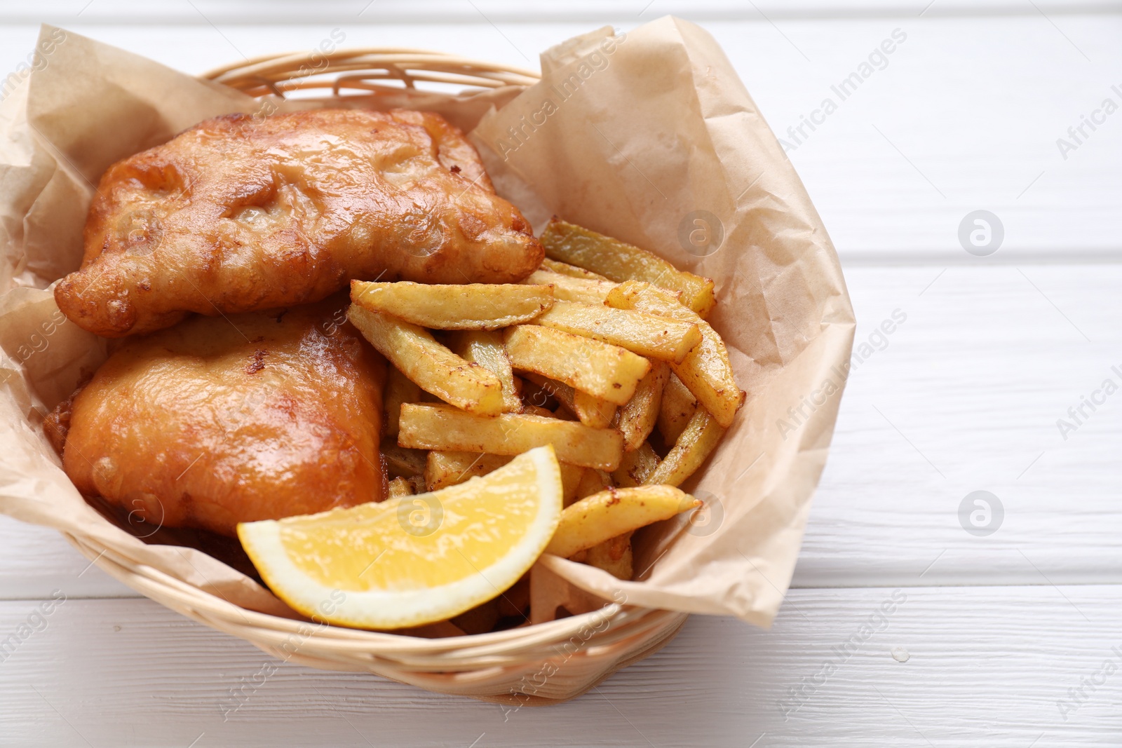 Photo of Tasty fish, chips and lemon in wicker bowl on white wooden table, closeup