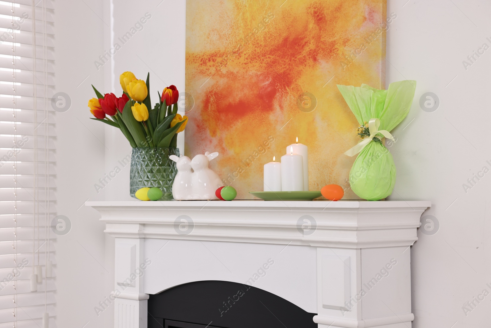 Photo of Easter decorations. Bouquet of tulips in vase, burning candles and bunny figures on fireplace at home