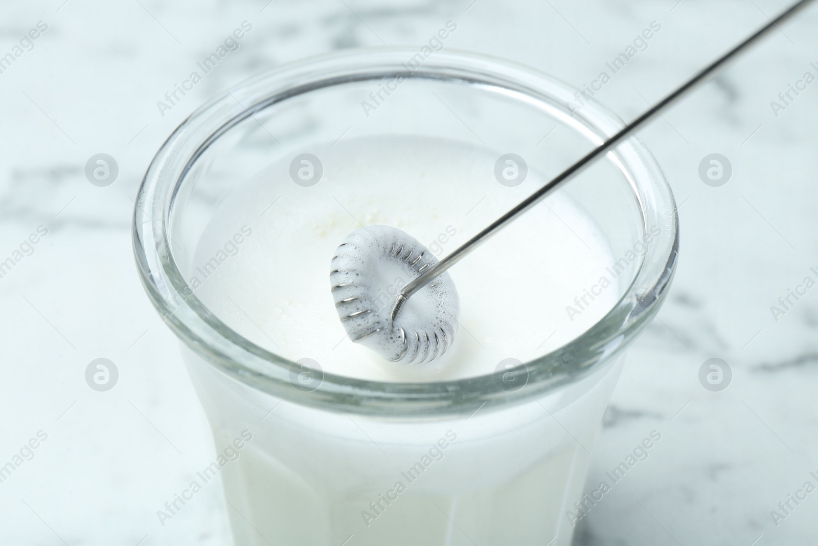 Photo of Whisking milk in glass with mini mixer (milk frother) at white marble table, closeup