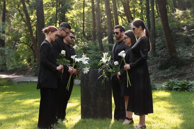 Photo of Sad people with flowers mourning near granite tombstone at cemetery outdoors. Funeral ceremony