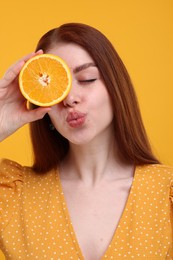 Beautiful woman covering eye with half of orange and sending air kiss on yellow background