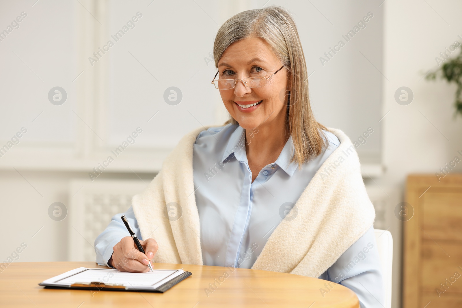 Photo of Smiling senior woman signing Last Will and Testament at table indoors