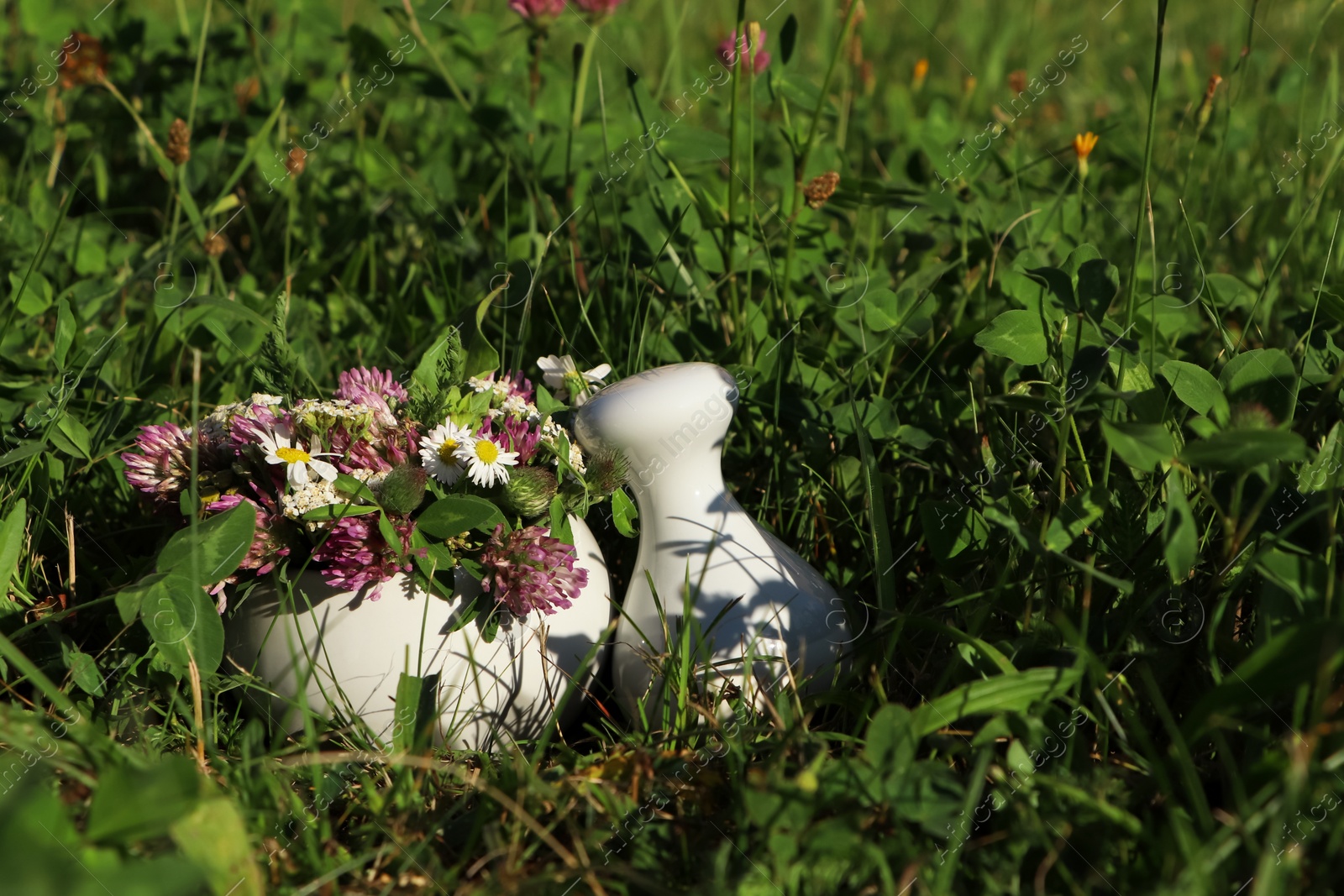 Photo of Ceramic mortar with pestle, different wildflowers and herbs on green grass outdoors. Space for text