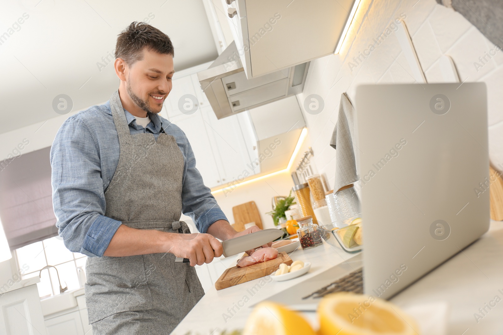 Photo of Man cutting chicken fillet while watching online cooking course via laptop in kitchen