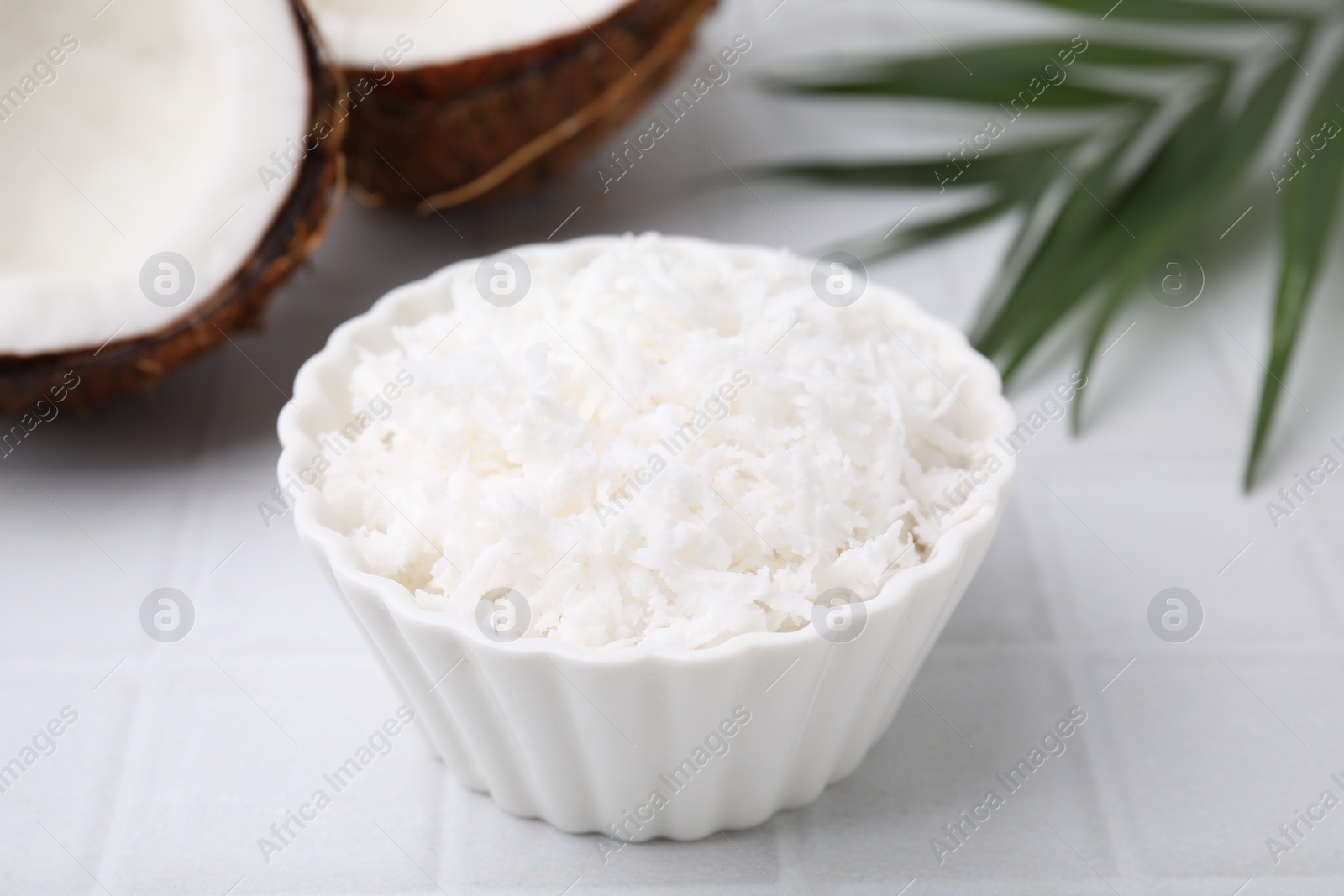 Photo of Coconut flakes in bowl, nuts and palm leaf on white tiled table, closeup