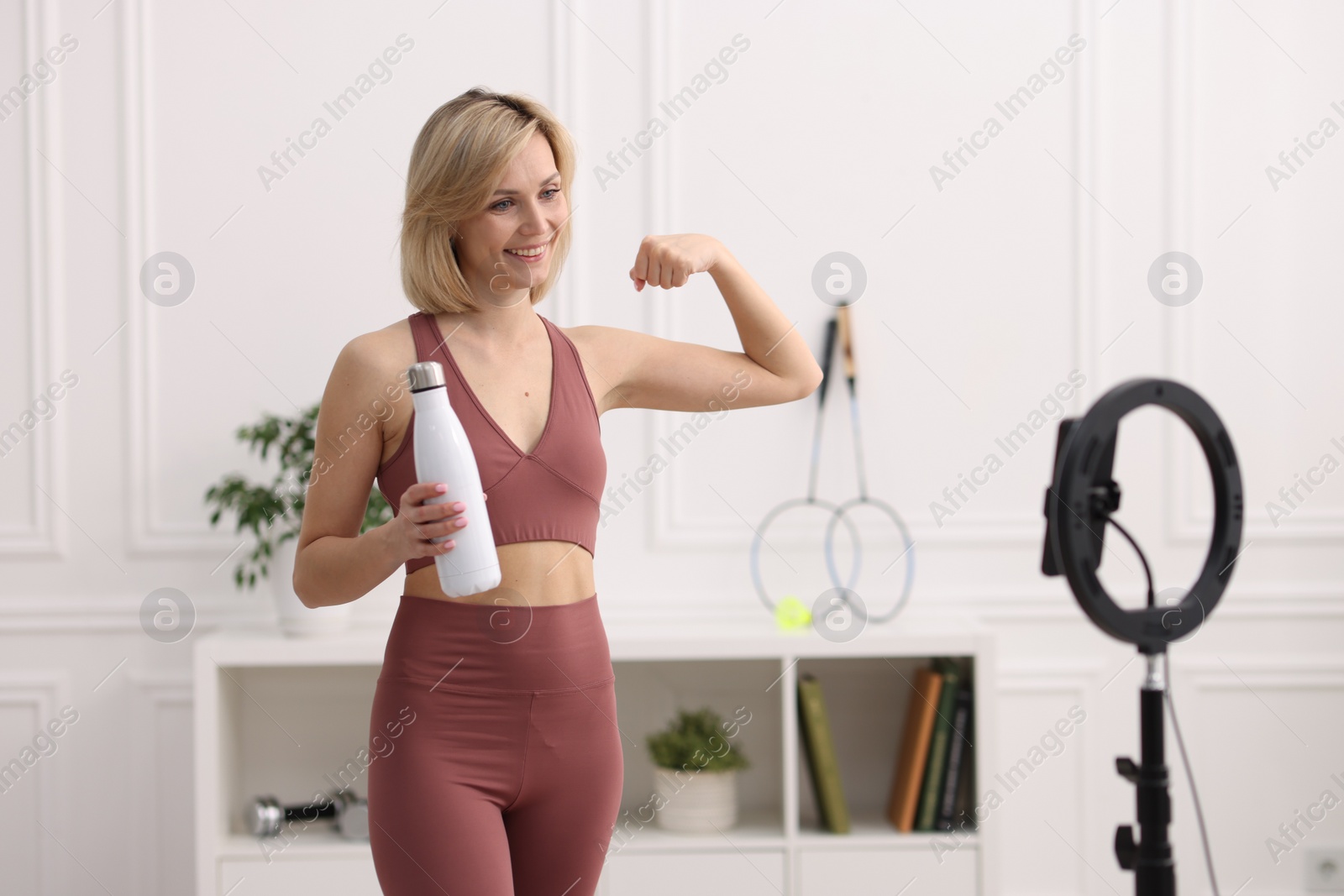 Photo of Smiling sports blogger holding bottle while streaming online fitness lesson at home