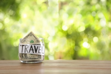 Glass jar with money and word TRAVEL on table against blurred background, space for text