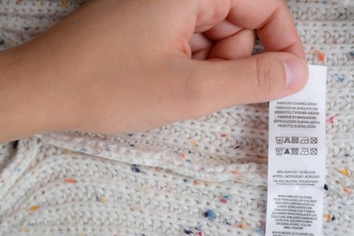 Photo of Woman holding white clothing label on beige warm sweater, closeup