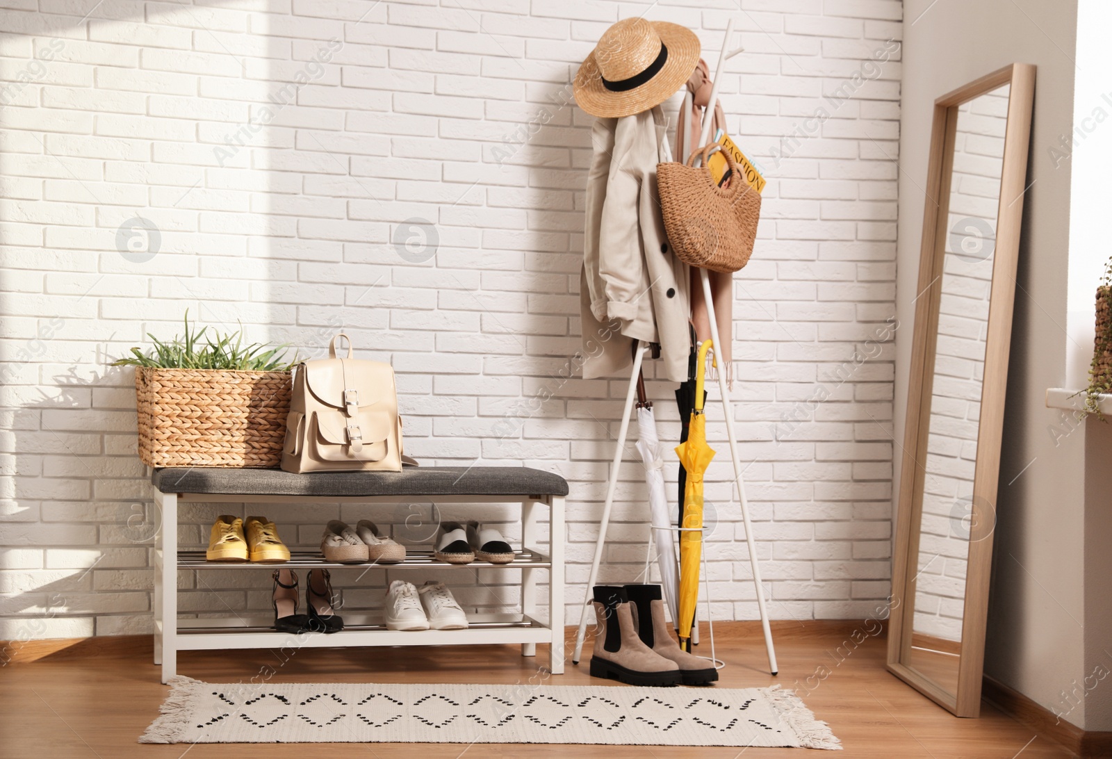 Photo of Stylish hallway interior with coat rack, shoe storage bench and mirror near white brick wall