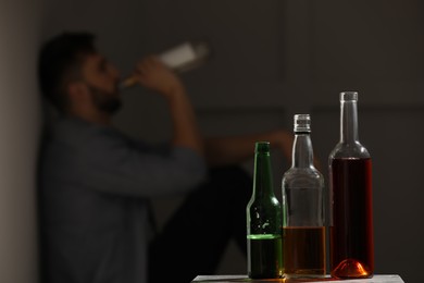 Addicted man sitting near wall indoors, focus on table with alcoholic drinks. Space for text