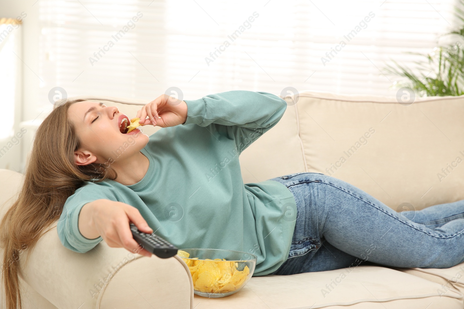 Photo of Lazy young woman with bowl of chips watching TV on sofa at home