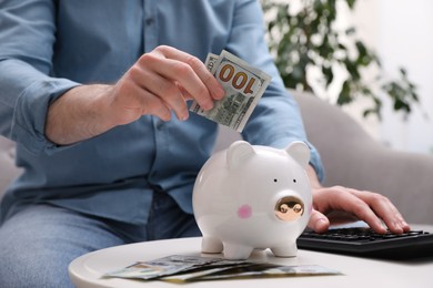 Man with calculator putting money into piggy bank at table indoors, closeup