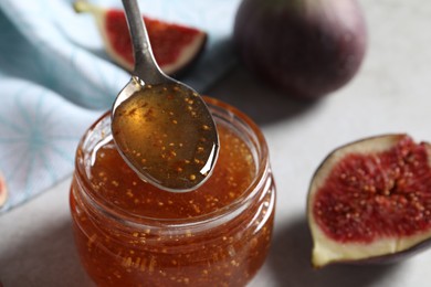 Photo of Spoon with tasty sweet jam over jar and fresh figs on light table, closeup