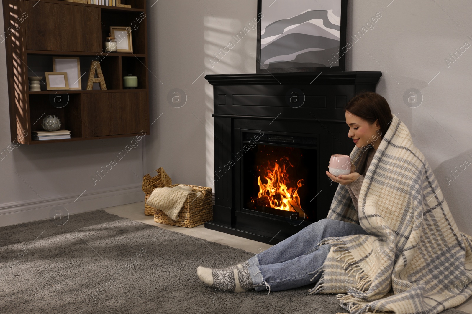 Photo of Beautiful young woman with cup of hot drink sitting on floor near fireplace at home. Space for text