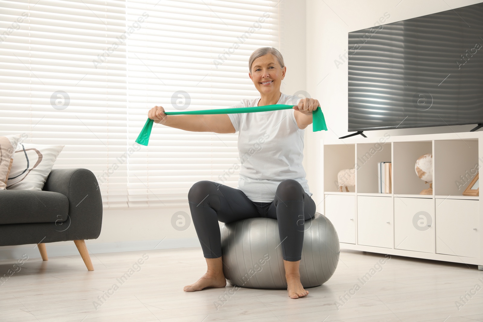 Photo of Senior woman doing exercise with elastic resistance band on fitness ball at home