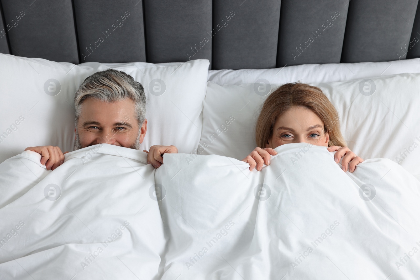 Photo of Lovely mature couple hiding together under blanket in bed at home, above view