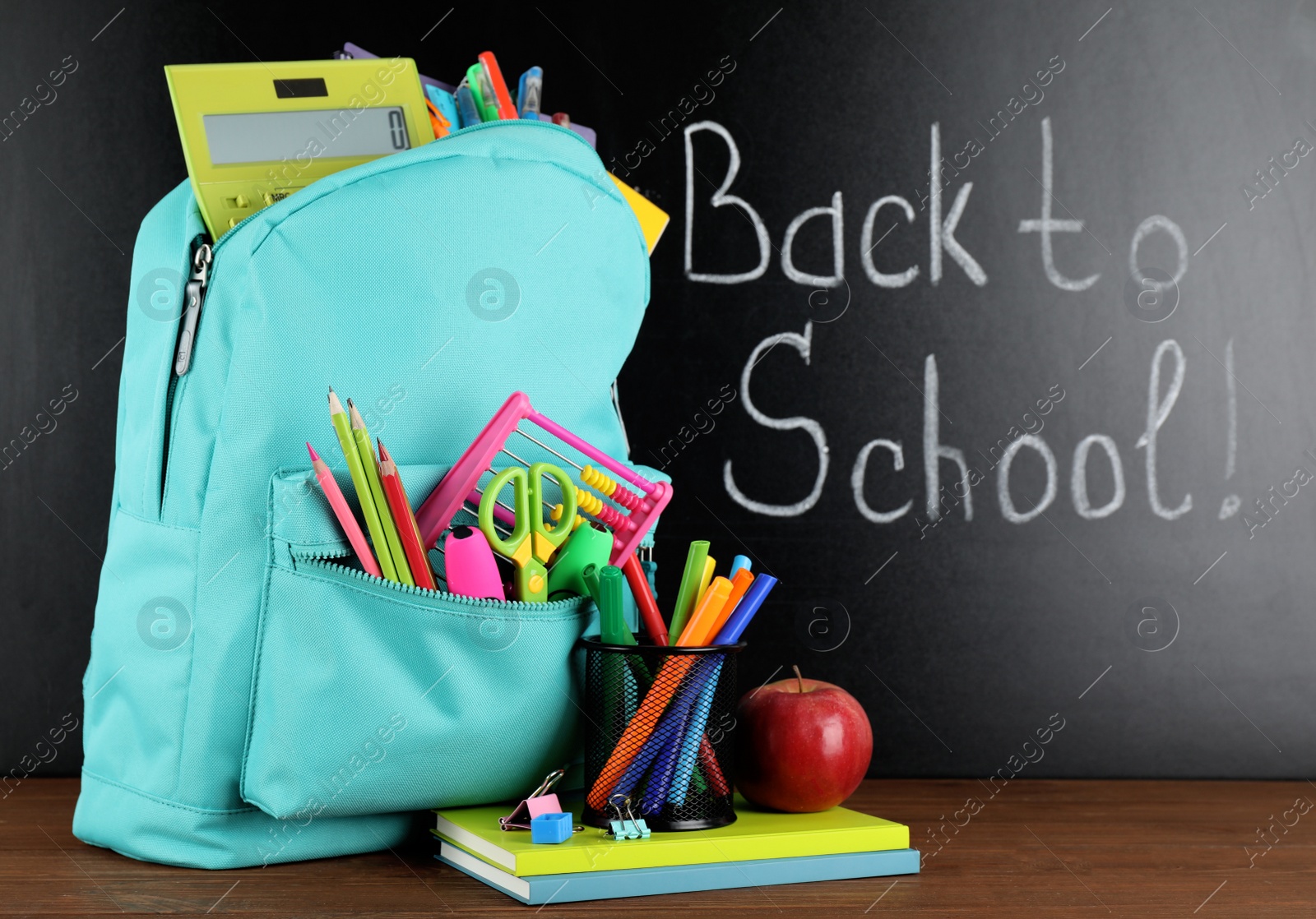 Photo of Bright backpack with school stationery on brown wooden table near black chalkboard. Back to School