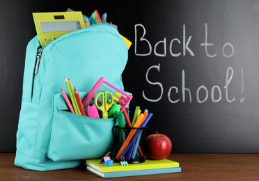 Photo of Bright backpack with school stationery on brown wooden table near black chalkboard. Back to School
