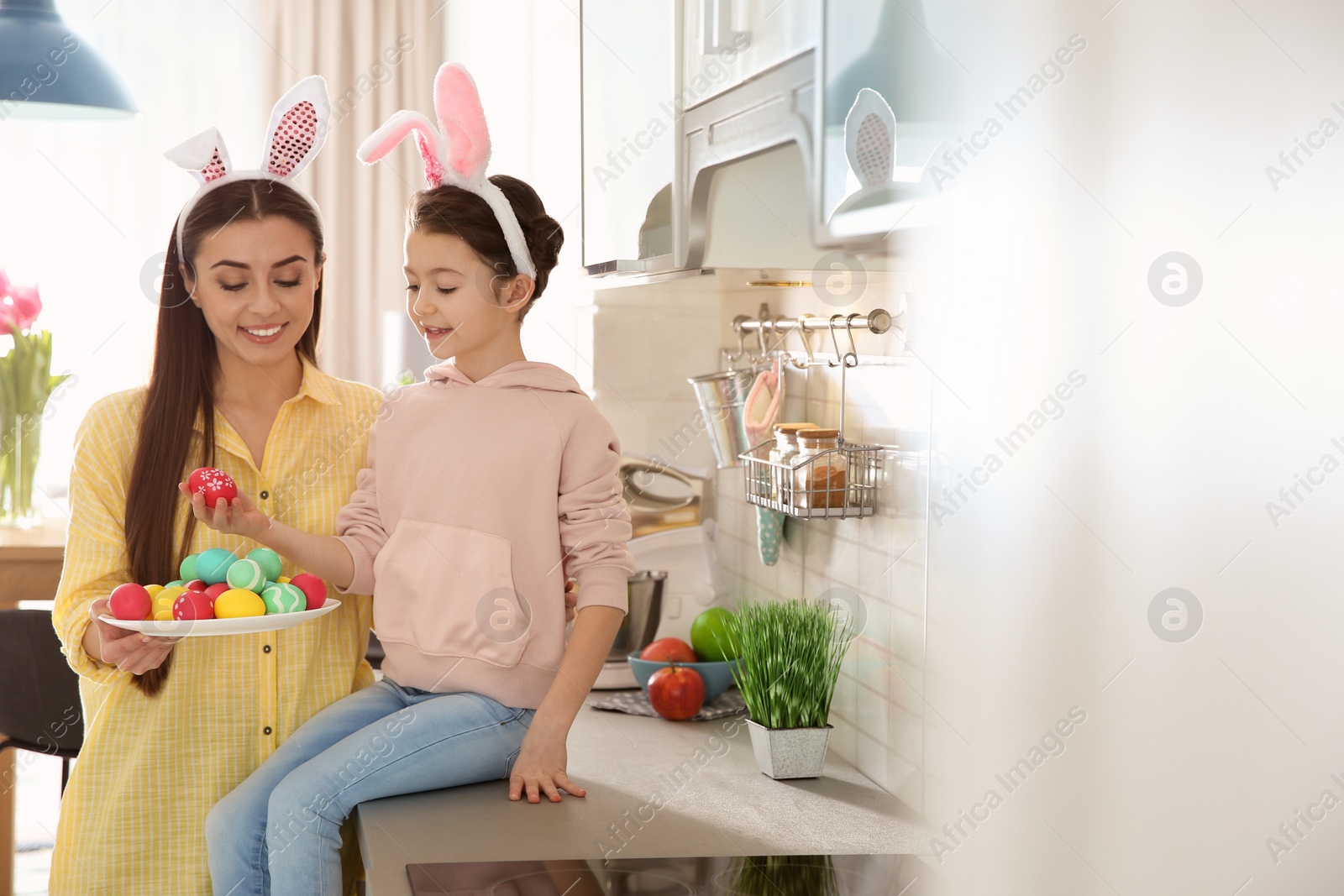 Photo of Mother and daughter with bunny ears headbands and painted Easter eggs in kitchen, space for text