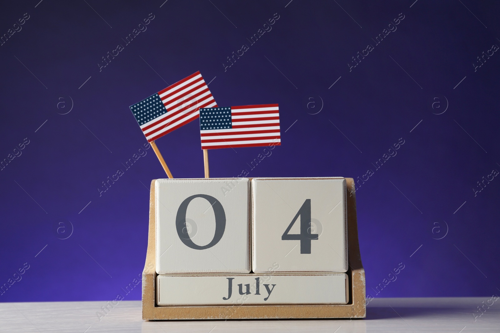 Photo of Wooden calendar with USA flags on table against color background. Happy Independence Day