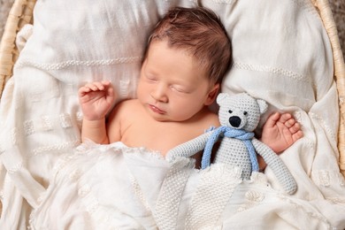 Cute newborn baby sleeping with toy bear on white blanket in wicker crib, top view