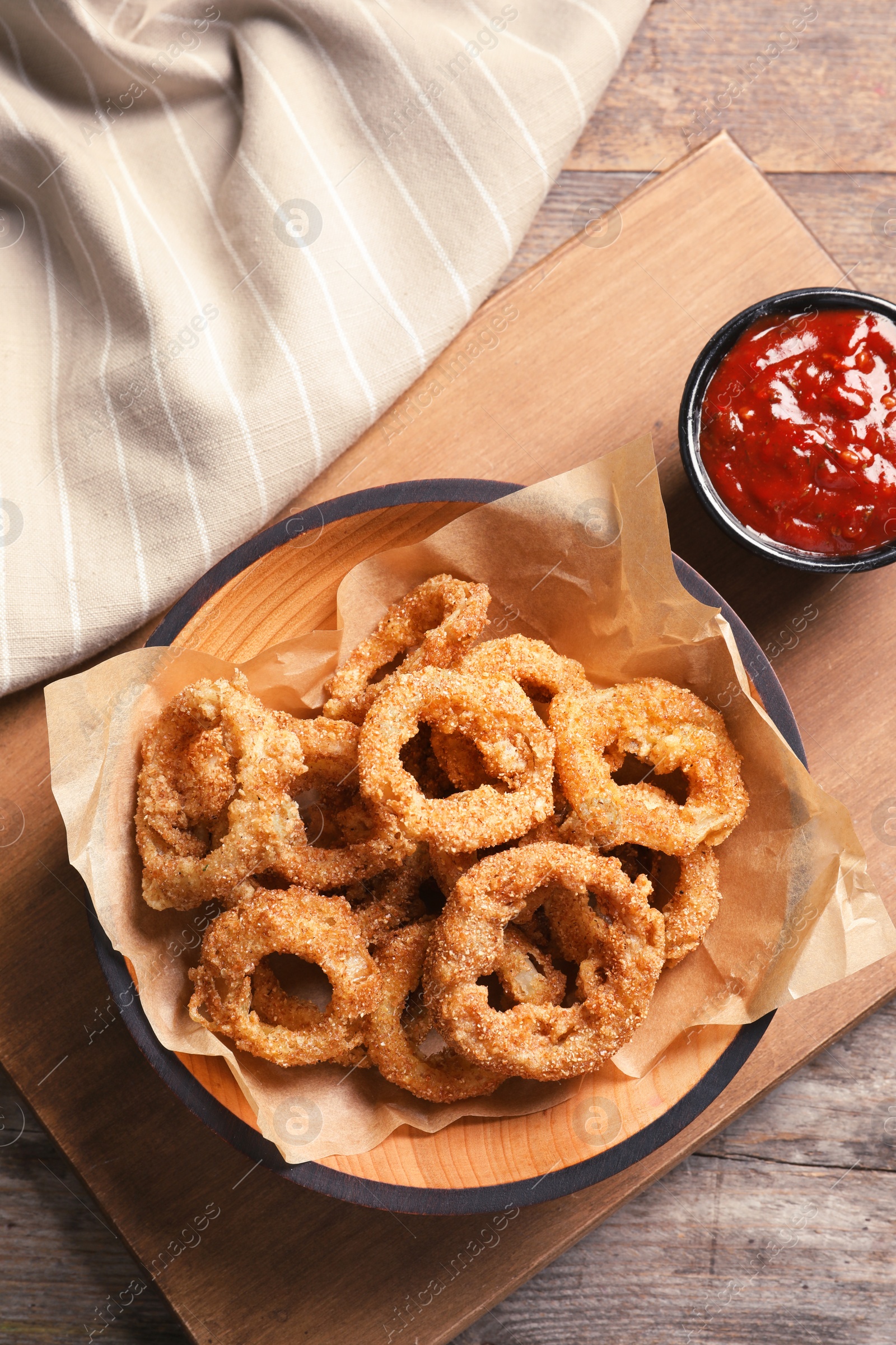 Photo of Homemade crunchy fried onion rings in plate and sauce on wooden background, top view