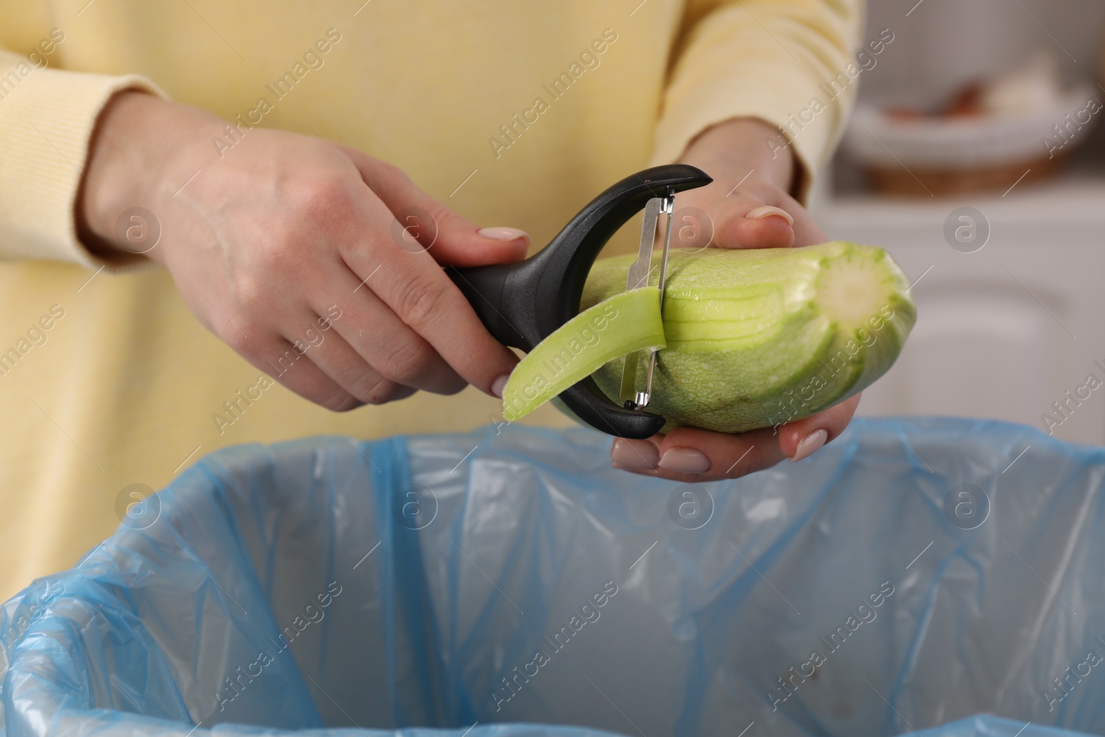 Photo of Woman peeling fresh zucchini above garbage bin indoors, closeup