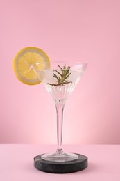 Photo of Martini glass of cocktail with lemon slice and rosemary on stand against pink background