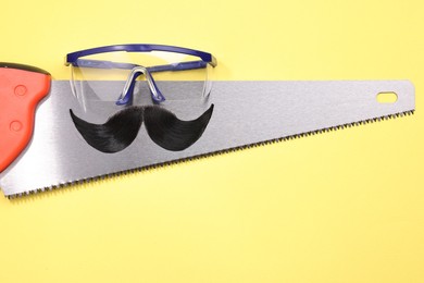 Photo of Man's face made of artificial mustache, safety glasses and hand saw on yellow background, top view. Space for text