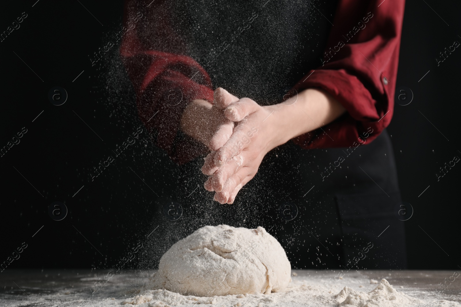 Photo of Making bread. Woman sprinkling flour over dough at table on dark background, closeup