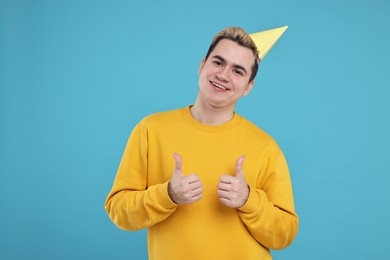 Photo of Young man with party hat showing thumbs up on light blue background