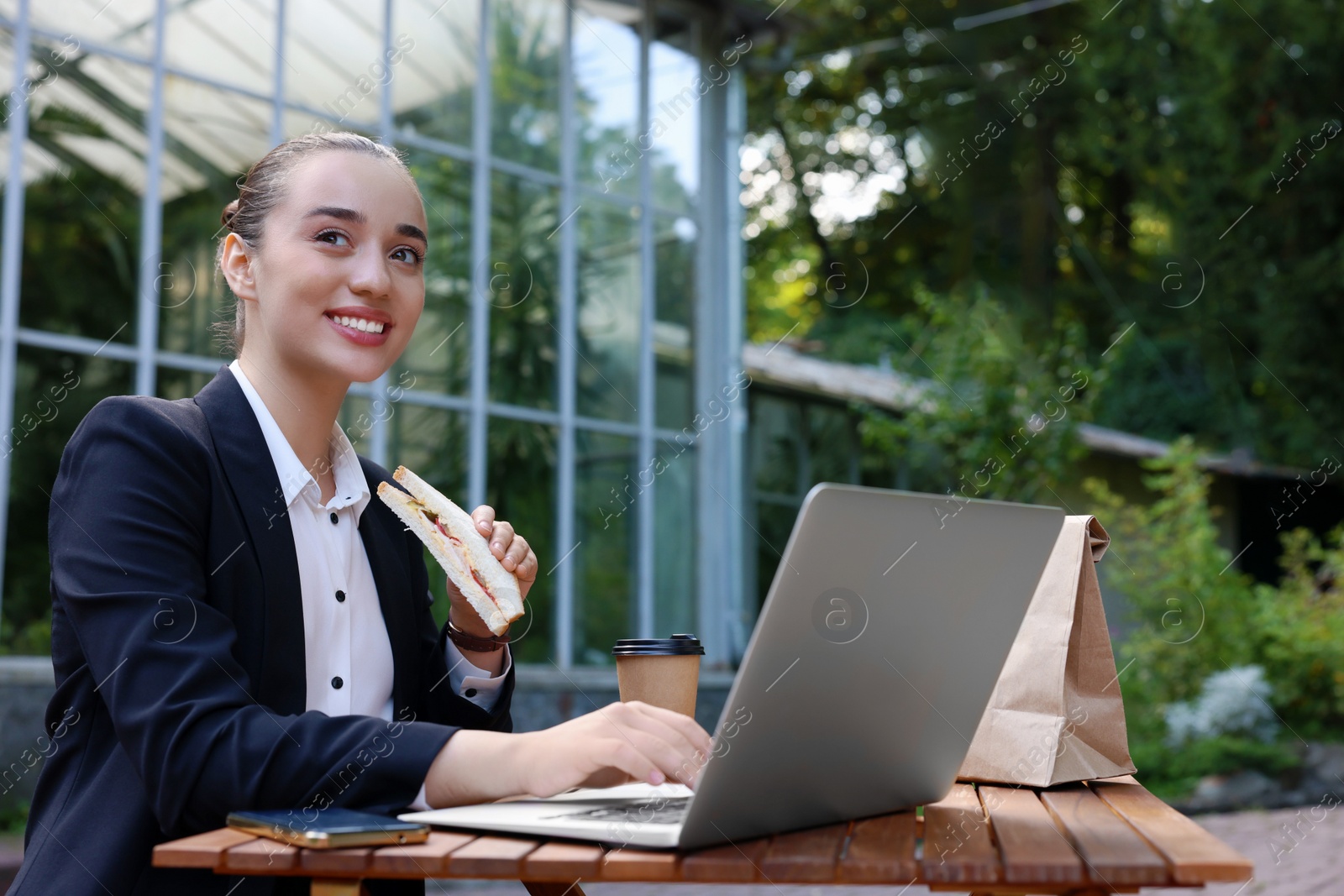 Photo of Happy businesswoman with sandwich using laptop while having lunch at wooden table outdoors