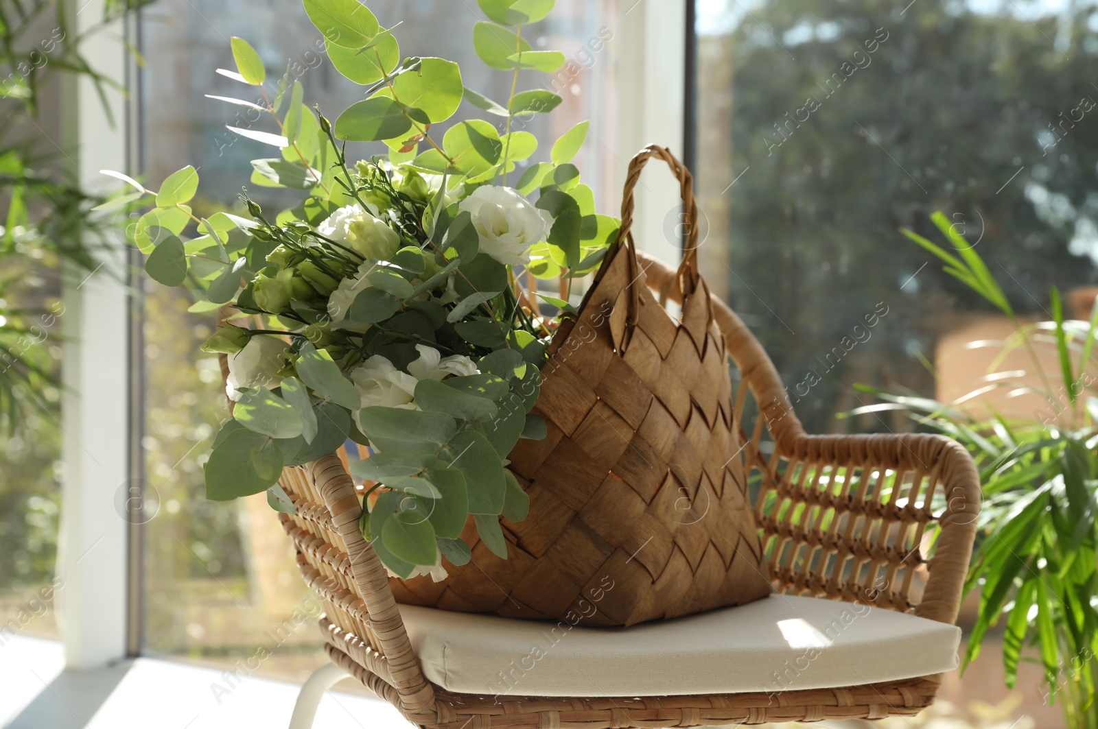 Photo of Stylish wicker basket with bouquet of flowers on chair near window indoors