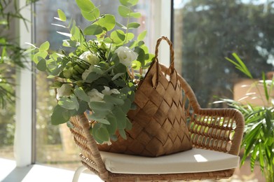 Stylish wicker basket with bouquet of flowers on chair near window indoors