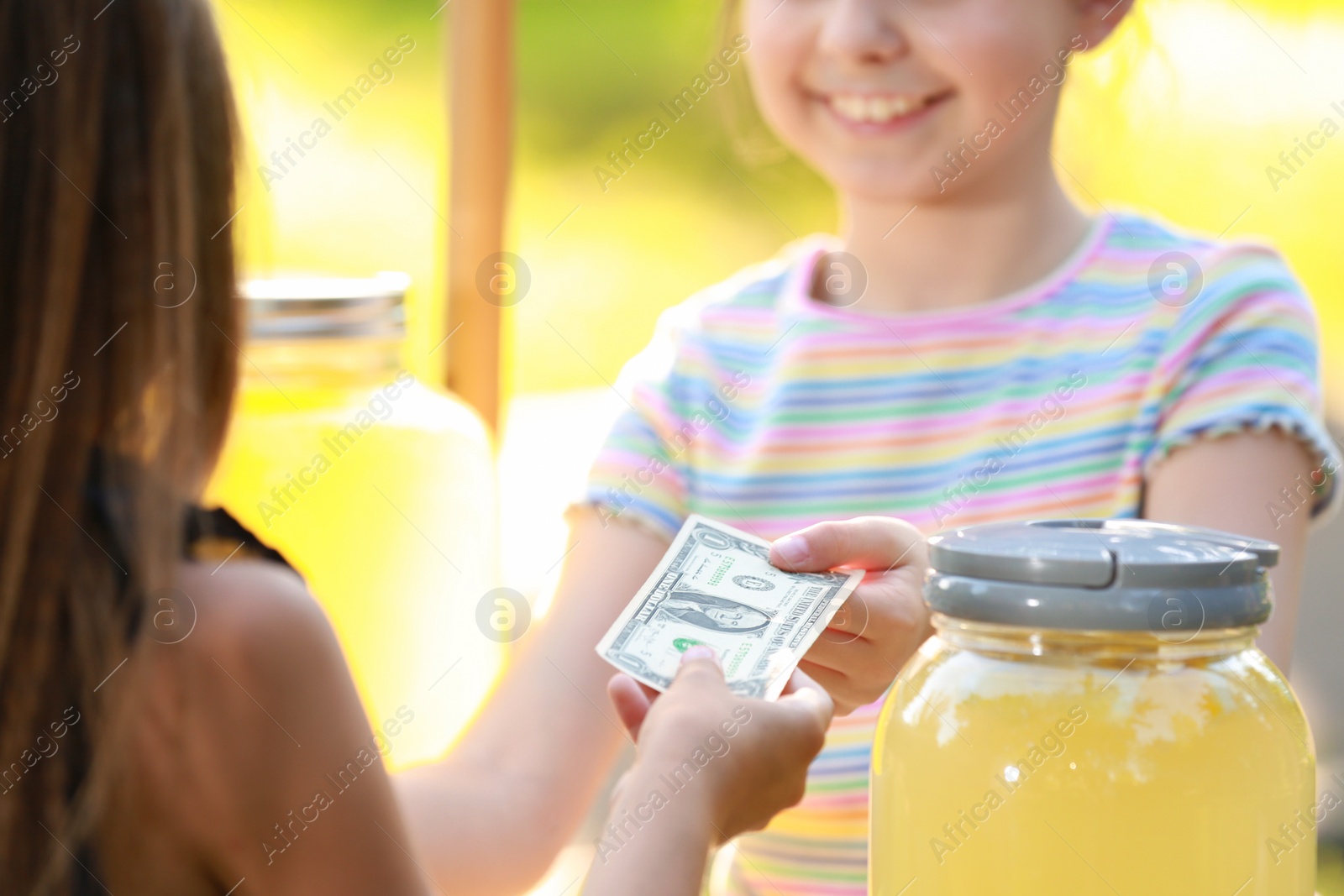 Photo of Little girl selling natural lemonade to kid in park, closeup. Summer refreshing drink