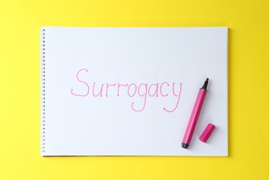 Photo of Notebook with word Surrogacy and marker on yellow background, top view